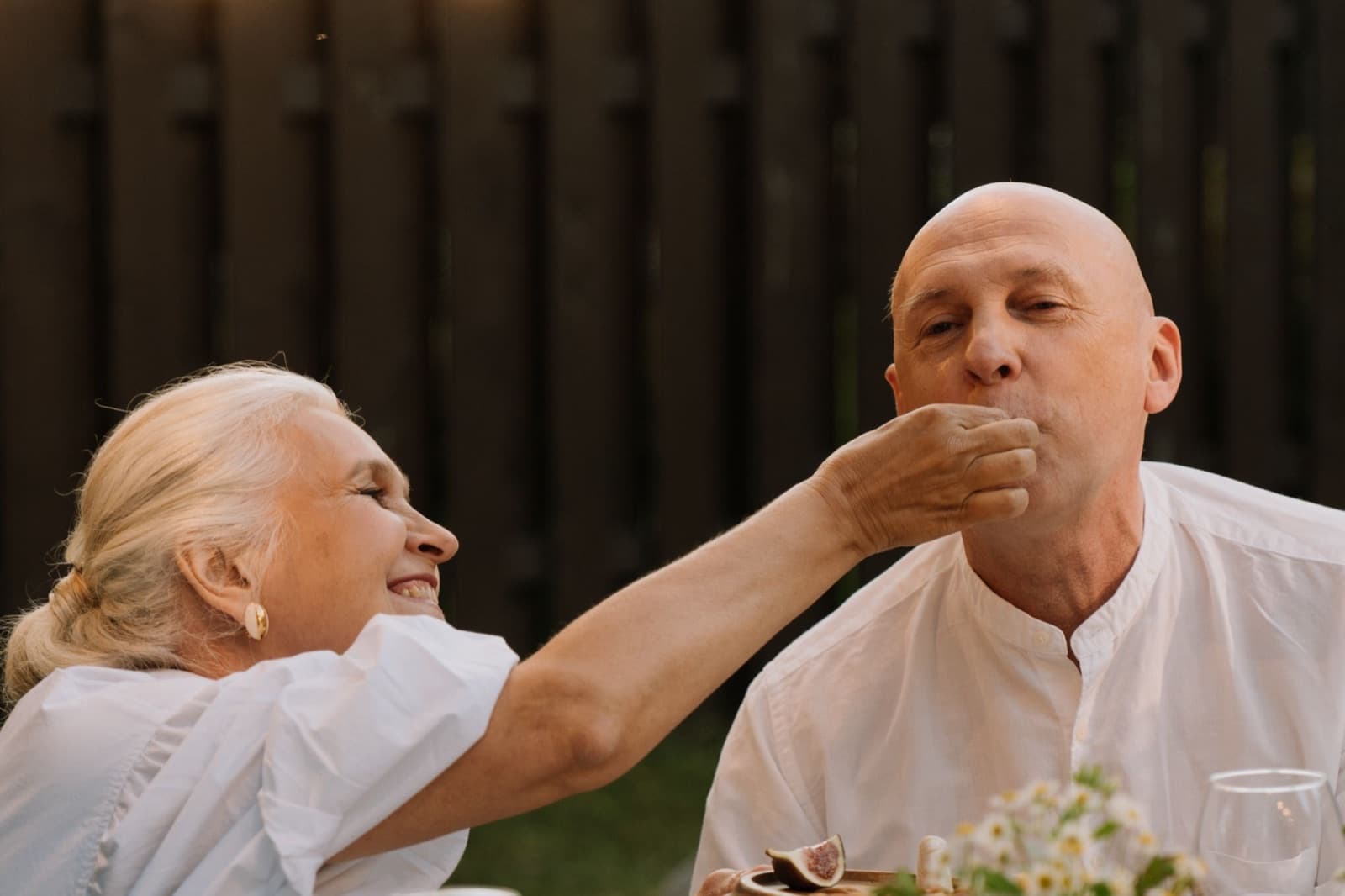 Een oudere vrouw laat haar man lachend een hapje proeven aan een gedekte tafel in de tuin