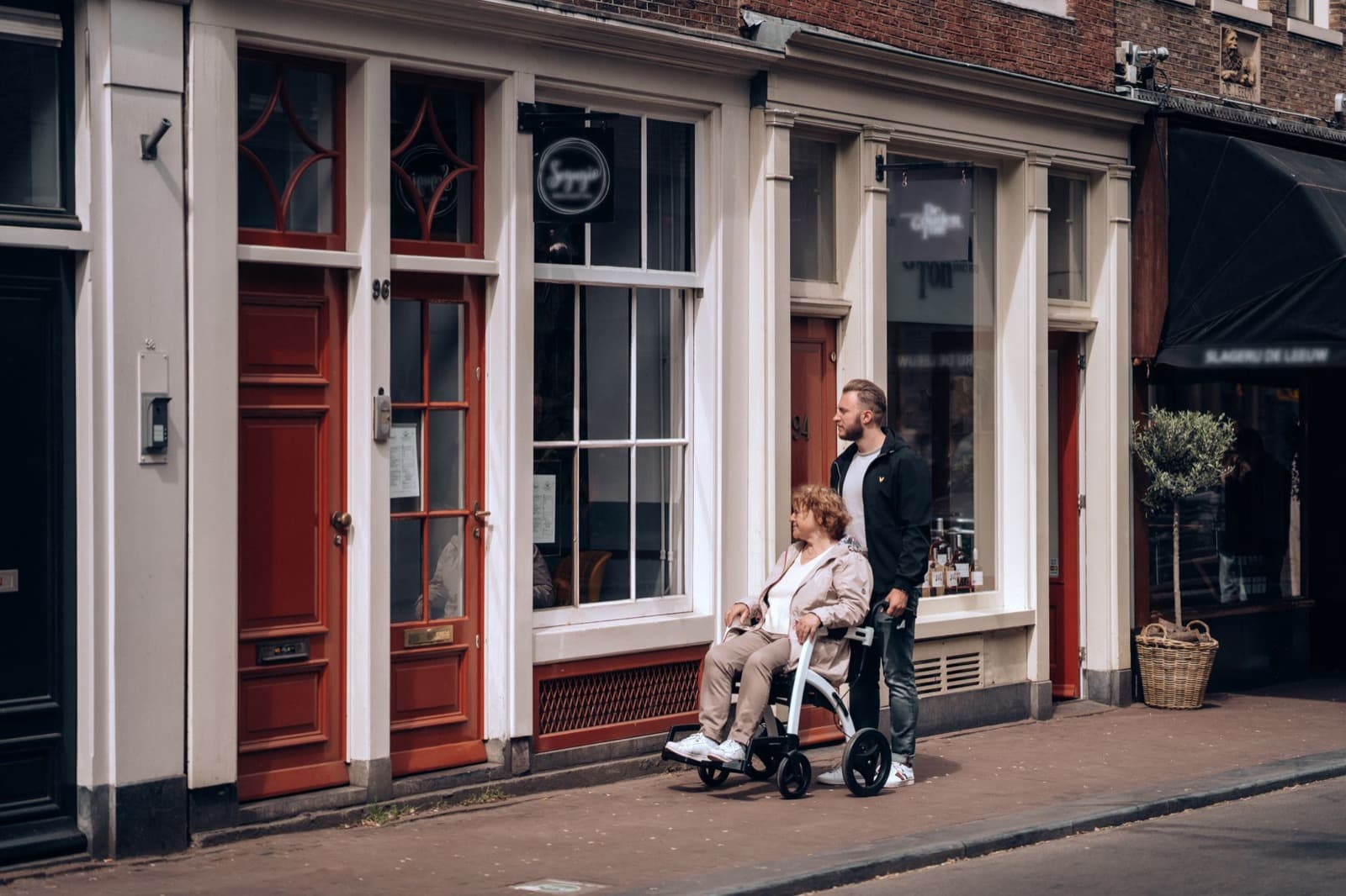 A man pushes a woman in a wheelchair through a Dutch street with brick houses